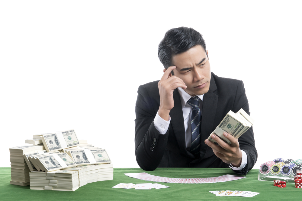 Asian man holding bundles of dollars and sitting at a poker table with cash laid on top