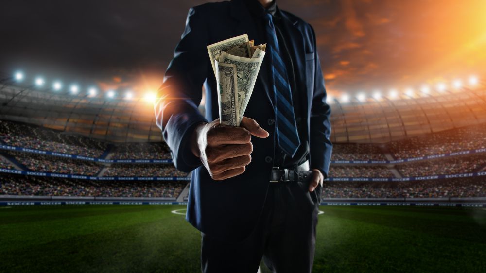 man in suit stands inside a stadium holding dollar notes in hand