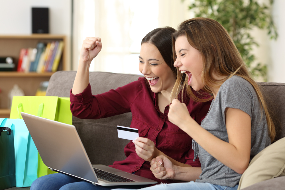 two young ladies sitting on a sofa cheering while looking at laptop and holding a credit hard