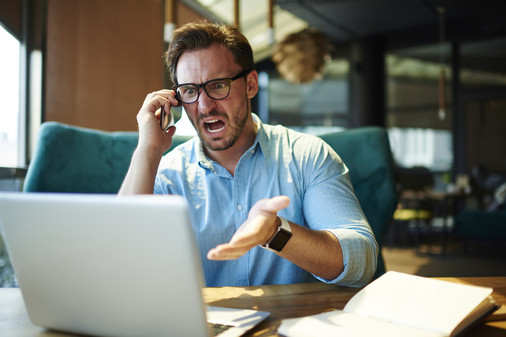angry man speaks on a cellphone while sitting at a desk in front of a laptop
