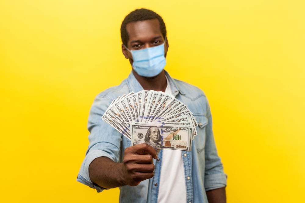 Man in surgical mask holds up American hundred-dollar bills