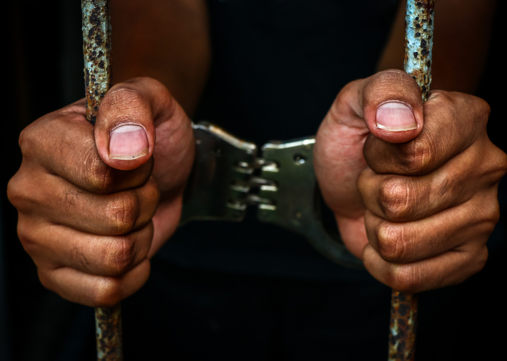 black man's hands in handcuffs and behind prison cell bars