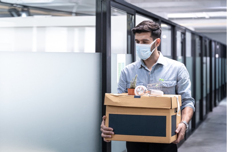 Man wearing a mask leaving an office with a box of belongings after losing his job