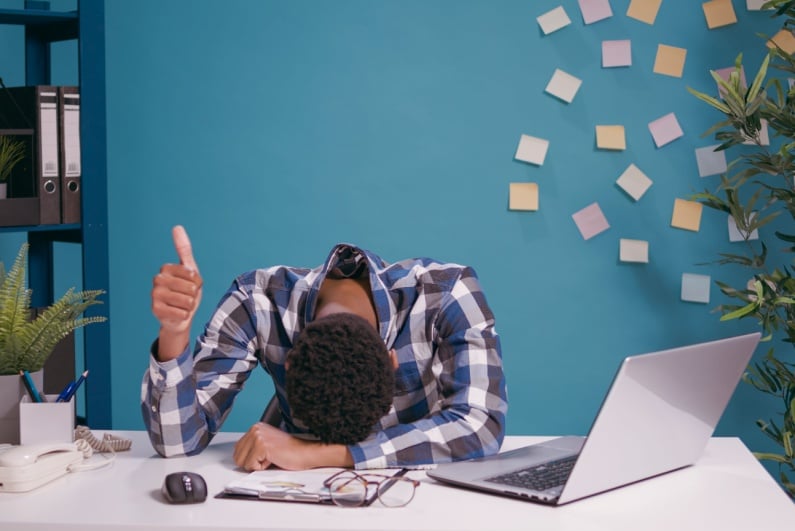 Tired man putting his head down on his desk and giving a thumbs up